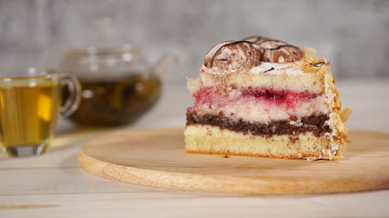 Close up of piece of cake on wooden board with green tea in teapot and cup on white wooden table.