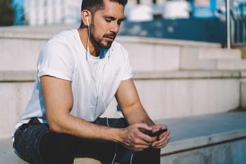 Serious caucasian bearded sportsman in earphones browsing new music via cellular for listening during training outdoors, young muscular male recording his results via app on modern smartphone