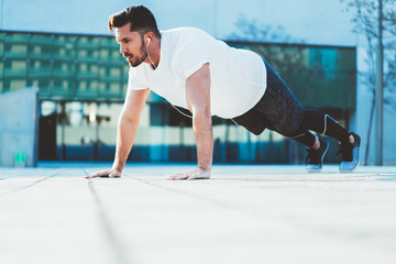 Fototapeta premium Young guy doing pushing from the floor outdoors training flexibility and strength reaching vitality goals, sportsman having morning workout doing exercises in earphones having physical power