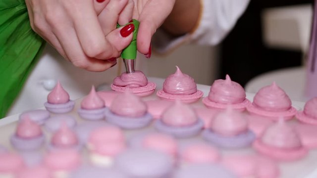 A smiling woman baker is making macarons with a pastry bag. The woman is putting filling for the tasty home-made macarons.