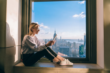Carefree digital nomad reading social publication with positive content text connecting to 4g wireless on modern touch pad, Caucasian blogger working remotely in highrise with lookout on Manhattan