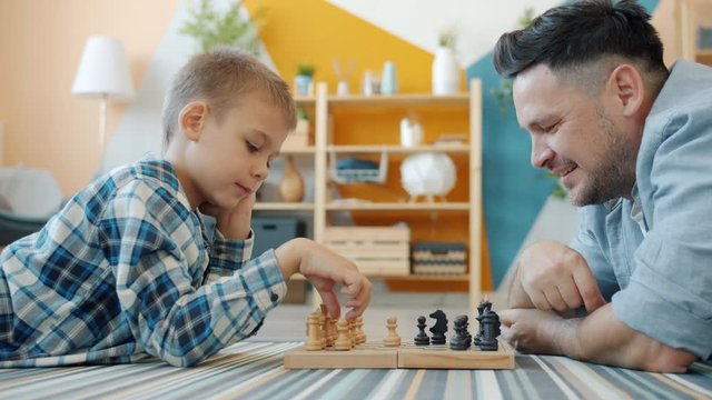 Smart child playing chess with dad at home lying on floor together having fun with intellectual sports game. Happy family and leisure activity concept.
