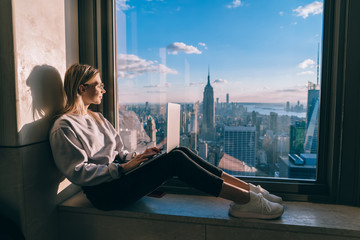 Young freelancer with laptop technology sitting at panoramic windowsill and enjoying beautiful above view of Manhattan district, millennial female with netbook working remotely in New York