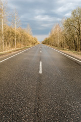 Straight wet asphalt road after a rain. On the sidelines are trees and bushes without foliage