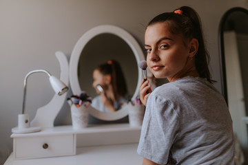 A young beautiful caucasian girl with a powder brush in front of a mirror