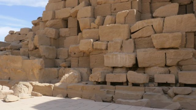 Lots of Stones of Brown and Yellow color Lie on the Ground. The Stone Wall Remains Are Stacked in a Row. The Stele with the Obelisk on the Top is Seen From Behind the Pile of Rocks.