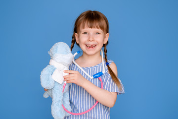 Smiling little girl playing a doctor and listening to a teddy bear with a stethoscope isolated on a blue background. Veterinary clinic game. The concept of the future profession.