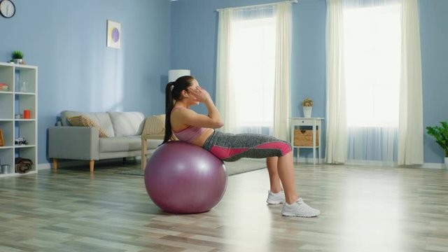 Young woman is training at home, doing upper crunches, using stability ball, listens her favorite sport music list, doing her best to get fit until summer time, vacation.