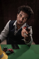 Portrait of young brunette Indian dark skinned man in vintage retro suit playing cards on a casino poker table in front of a textured copy space studio background. lifestyle and fashion.