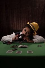Young Indian Bengali brunette woman in vintage western suits with yellow hat playing cards on a casino poker table in brown textured copy space studio background. Indian lifestyle and fashion.
