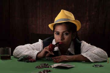 Young Indian Bengali brunette woman in vintage western suits with yellow hat playing cards on a casino poker table in brown textured copy space studio background. Indian lifestyle and fashion.