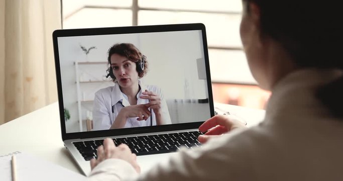 Videoconferencing doctor concept. Female therapist wears headset providing remote medical assistance in virtual conference chat consulting patient online by webcam on laptop screen. Over shoulder view