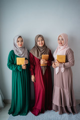 three moslem women smilling hold the holy book of Al-Quran when standing on the wall background