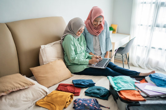two muslim woman using laptop to looking departure schedules via online application after preparing and put her clothes in the suitcase