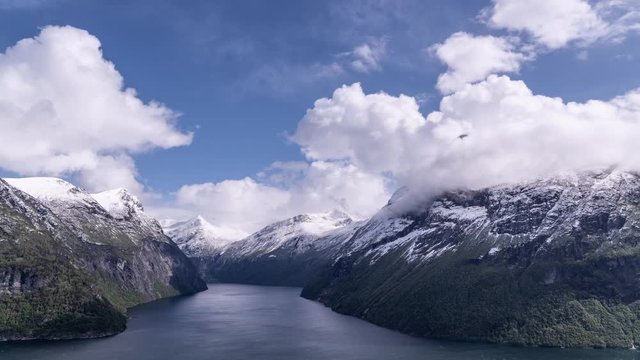 Beautiful white clouds whirling above the calm blue water of the Geiranger fjord. Steep cliffsides covered with scarce vegetation. Fresh snow laying on the mountain tops. Blue skies above.