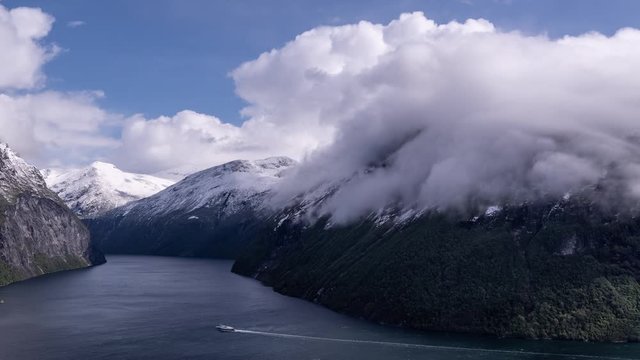 White clouds whirling above the calm blue water of the famous Geiranger fjord. Steep cliffsides covered with scarce vegetation. Fresh snow laying on the mountain tops. Blue skies above.