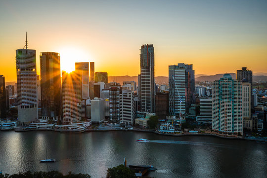 Brisbane City Skyline View With Sunset Sun Streaking Light Beams With Boat Crossing The River