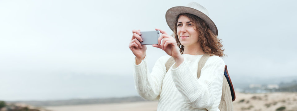 Handsome woman with hat and backpack taking photo with her smartphone, traveling among natural sand meadows