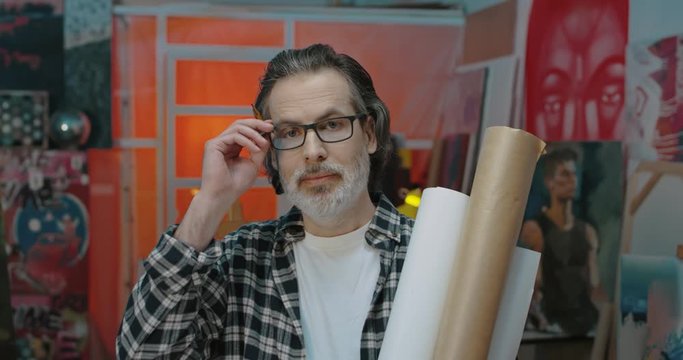 Portrait of handsome middle-aged Caucasian man in glasses and with gray beard standing in workshop, holding tubes of papers and looking at camera. Male artist in cozy messy studio.