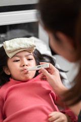 Asian mother measuring temperature girl with digital thermometer in her mouth on bed at morning time, Sick child have cool towel for reduce high fever,  Selective focus, Healthy and infection concept