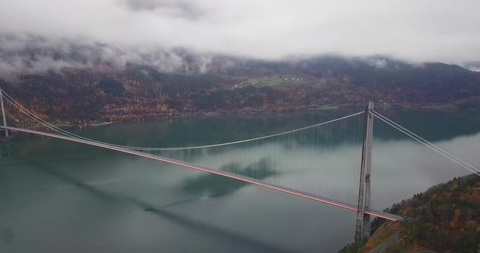 An aerial view of the Hardanger Bridge. The suspension bridge spanning above the calm water of the Eidfjord, thick white clouds hanging above, dense forest growing on both sides of the fjord.
