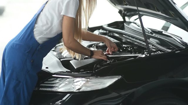 A woman car mechanic working in a garage and examining a car. Repair service.