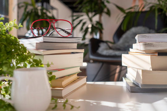 Inside a green house with many plants. A pile of different books on a with table with red glasses and a cup of coffee at sunset