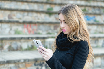 Stylish Young blonde woman looking at her smart phone checking social media and e-mail accounts to connect with friends and family outdoors