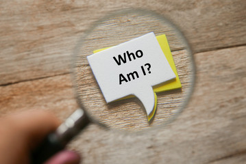Selective focus of hand holding magnifying glass focusing a stack of speech bubbles written with question Who Am I? on a wooden background.