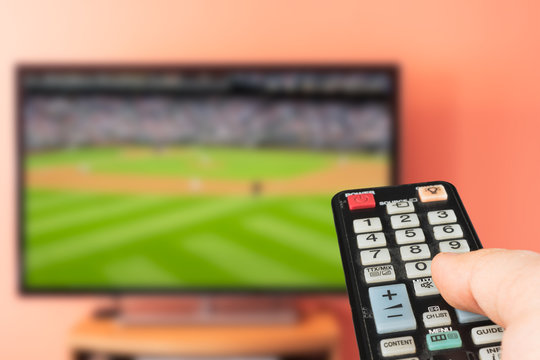 Caucasian man watching baseball game on TV at home. Changing channels and adjusting volume with television remote control. 