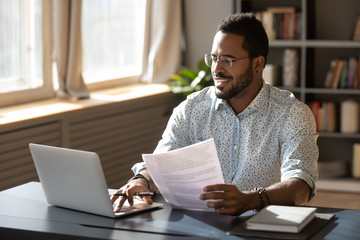 Happy millennial african american businessman in eyewear holding documents, doing paperwork, preparing report or analyzing market research results, working on computer in modern workplace office. - Powered by Adobe