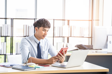 handsome asian male worker using mobile phone contacting colleagues, siting at desk with office equipment and devices such as laptop computer, books and pens, representing contact us in work area