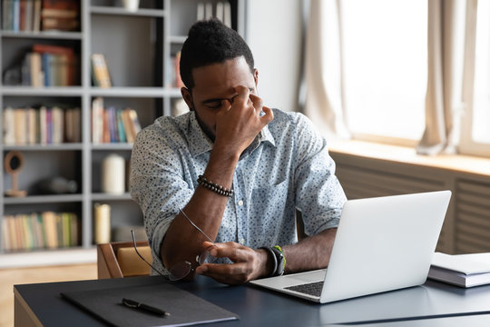 Frustrated Young Biracial Guy Taking Off Eyeglasses, Massaging Nose Bridge, Feeling Tired Due To Computer Overwork. Overwhelmed Stressed Millennial African American Man Suffering From Eyes Strain.