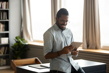 Happy young handsome mixed race man in eyewear leaning on table, looking at digital tablet screen,...