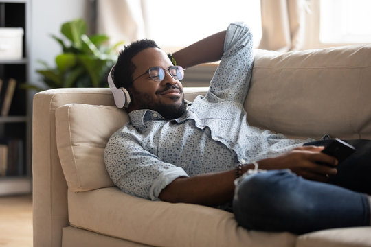 Tranquil carefree young african american man lying on comfy couch, wearing modern wireless headphones, listening to favorite classic music online, feeling peaceful mindful alone in living room.