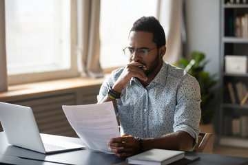 Thoughtful young mixed race businessman in glasses holding paper document, reading banking debt notification. Pensive millennial african american accountant employee reviewing research report.