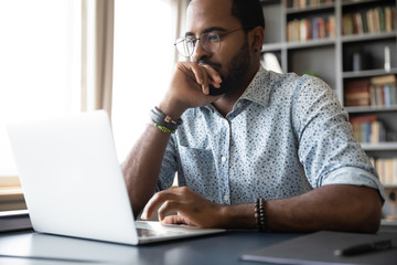 Thoughtful millennial biracial man in eyeglasses stack with hard task, looking at laptop screen. Puzzled young african ethnic businessman thinking of problem solution, making difficult decision. - Powered by Adobe