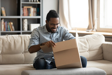 Happy young african ethnicity male client sitting on sofa at home, opening huge carton box,...