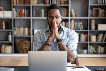 Fototapeta premium Front view young puzzled african american businessman in glasses looking in computer monitor, feeling doubtful about difficult business decision, sitting alone at table desk in office or home.