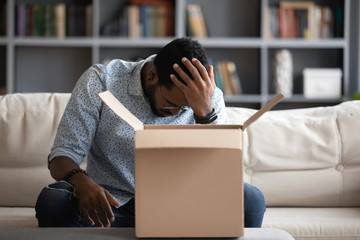 Stressed young multiracial man sitting on couch at home, feeling desperate about ordering wrong...