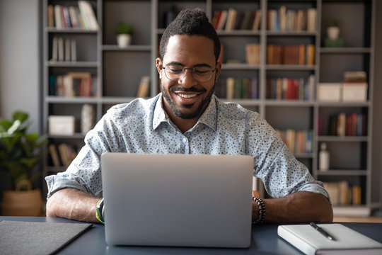 Front view head shot young smiling african american businessman in eyewear looking at laptop screen, reading pleasant messages in social networks, working on computer sitting at table in office home.