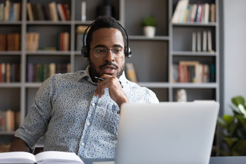 Focused millennial mixed race guy in glasses wearing wireless headset with microphone, looking at...