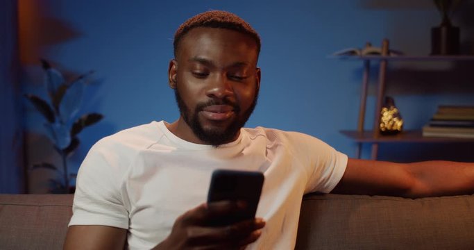 Front view of handsome african man looking at smartphone screen while sitting on couch. Bearded young guy chatting in social networks while spending free time at home