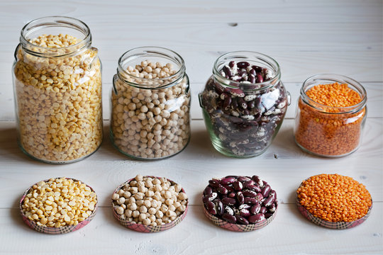 Legumes (peas, nute, beans, red lentils) in glass jars of various shapes on a white wooden background