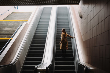 Beautiful young woman in trench coat stands on an escalator in the subway. - Powered by Adobe