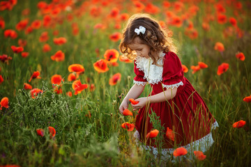 Cute happy girl with curly hair in a red vintage dress with lace in the rays of the setting sun on a poppy field.