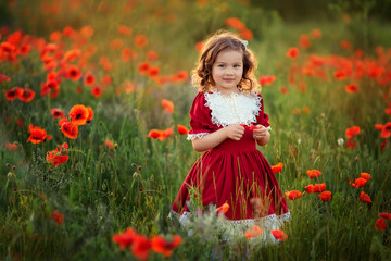 Naklejka na ściany i meble Cute happy girl with curly hair in a red vintage dress with lace in the rays of the setting sun on a poppy field.