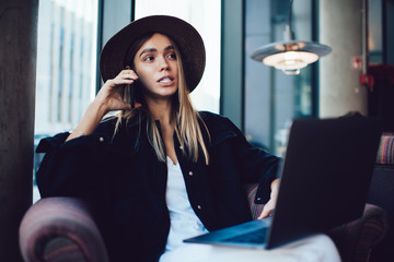 Stylish woman talking on smartphone while working on laptop in cafe