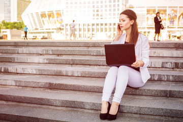 Beautiful brunette business woman in white suit with notebook on her lap, typing, working outdoors. Copy space