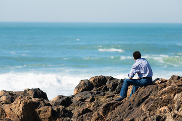 Fototapeta na wymiar Rear view of pensive, depressed man sitting on coast rock, looking at sea horizon.Copy space, unrecognizable person, lens flare, sunset light